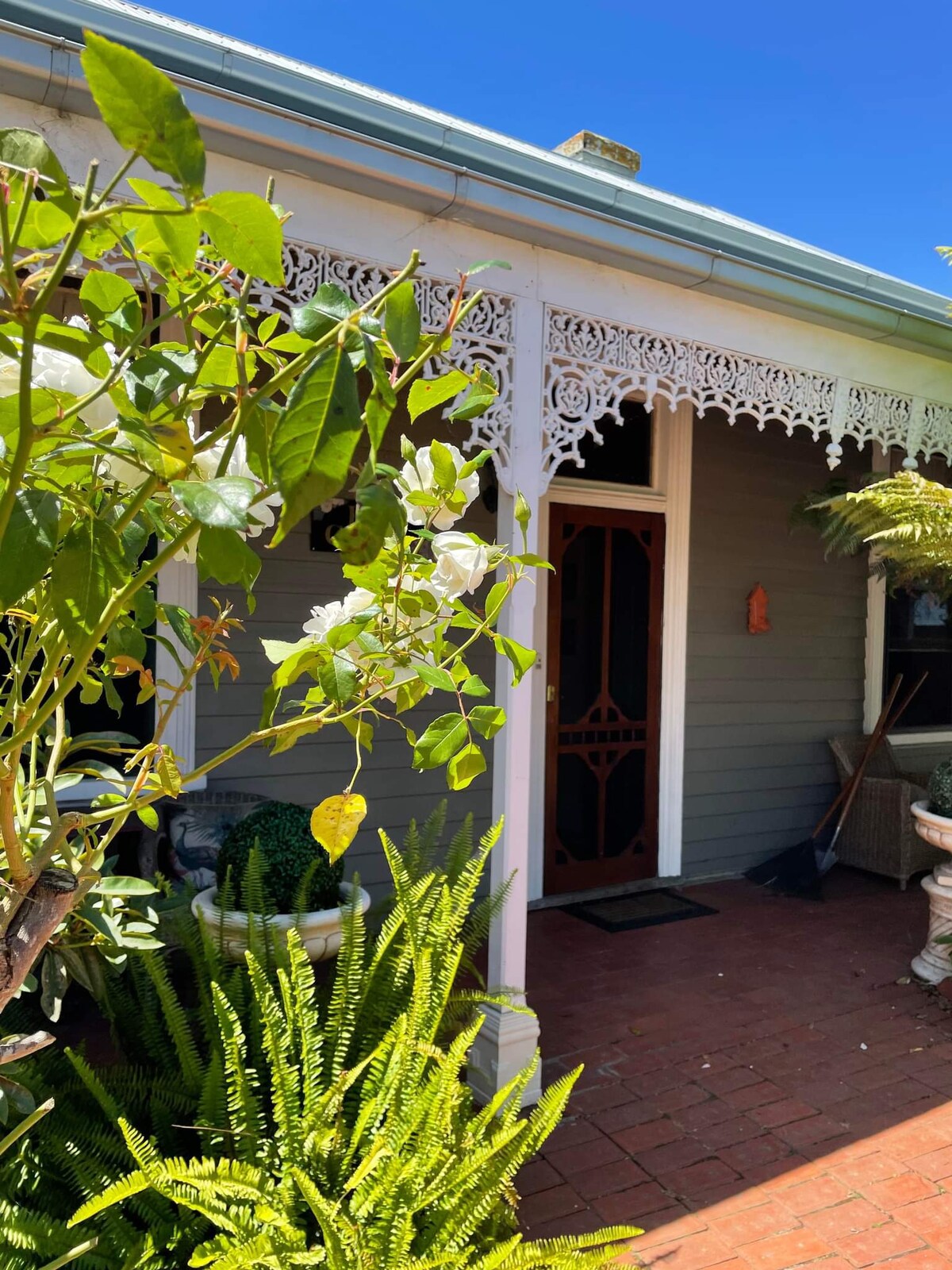 The entrance to the cottage features ornate Victorian detailing and a welcoming porch. Lush greenery, including ferns and flowering plants, surrounds the doorway, creating a calming atmosphere. A brick pathway leads to the intricately designed front door, set against a backdrop of clear blue sky.