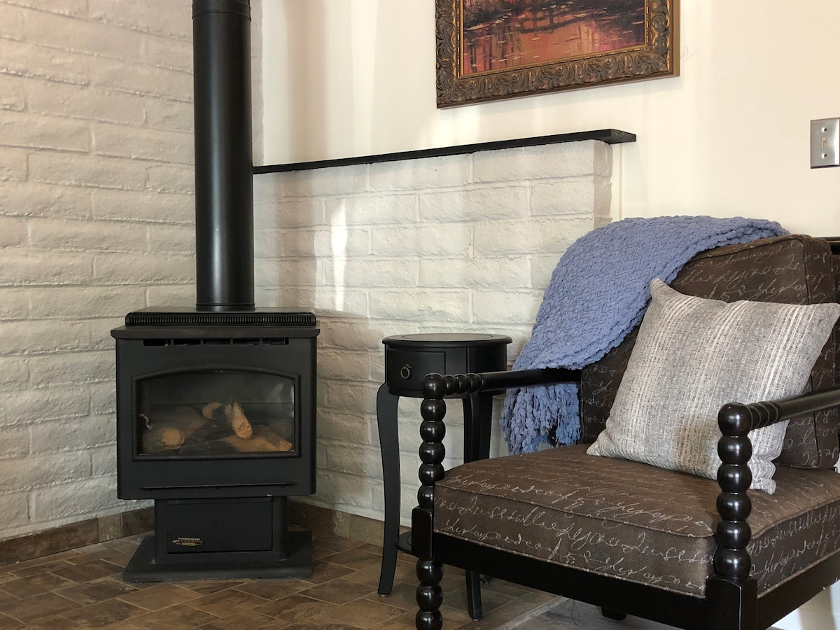 A comfortable seating area is highlighted by a dark, patterned chair and a small side table. A wood stove is positioned nearby, adding warmth to the space. Soft blue knit and a decorative pillow complement the inviting atmosphere.