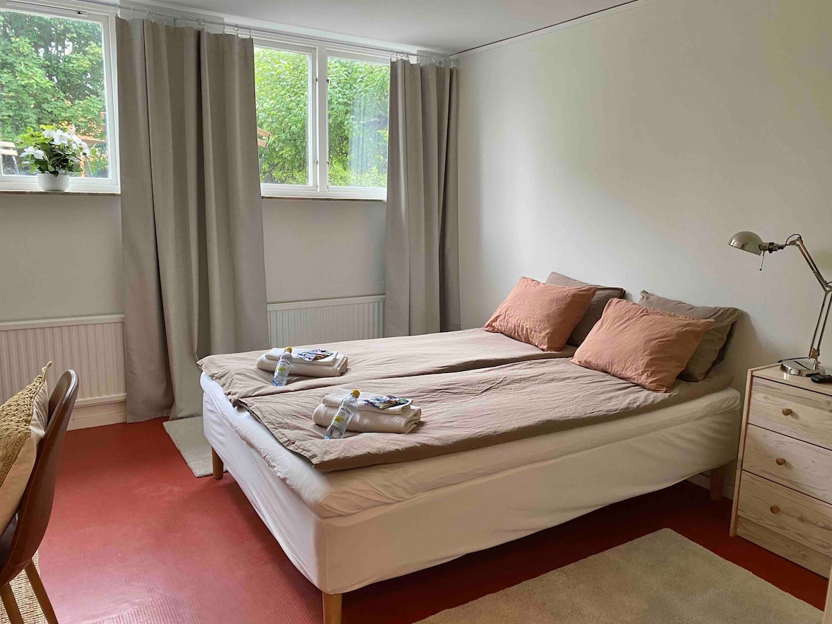 A well-lit room features a spacious bed adorned with neutral bedding and decorative pillows. A small bedside lamp sits next to the bed, and a wooden dresser is positioned nearby. Floor-to-ceiling curtains frame the window, allowing natural light to brighten the space.