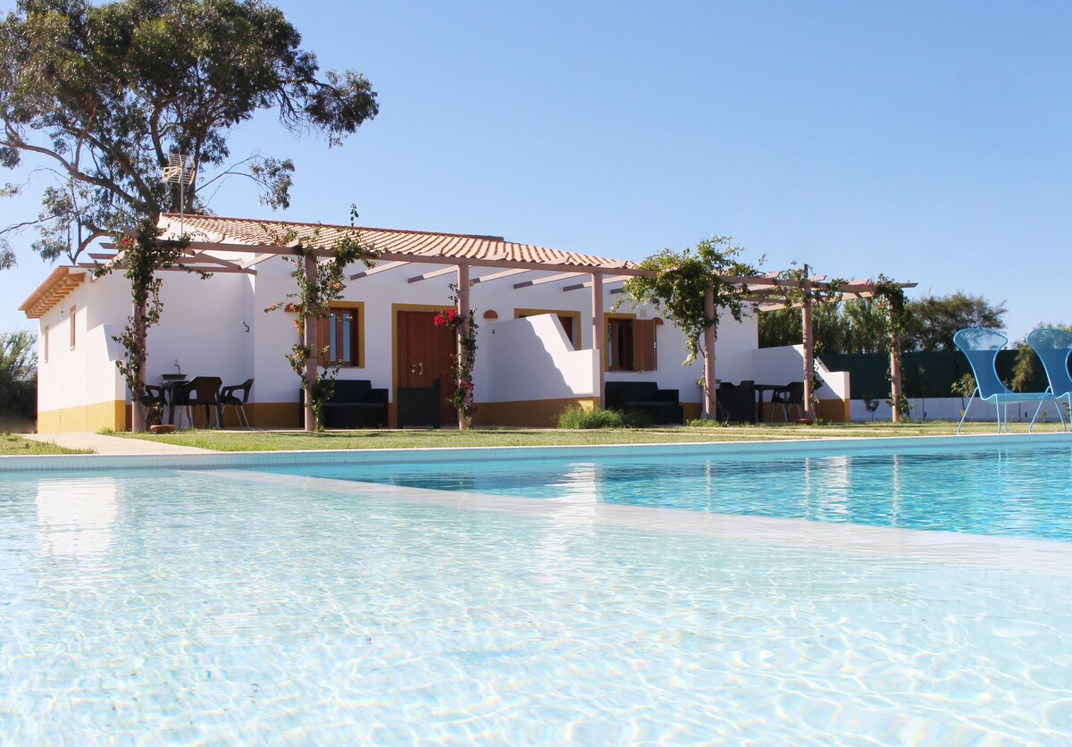 The image depicts a row of three charming white houses with terracotta roofs set against a clear blue sky. A tranquil swimming pool in the foreground reflects the sun, while lush greenery and climbing vines enhance the outdoor ambiance.