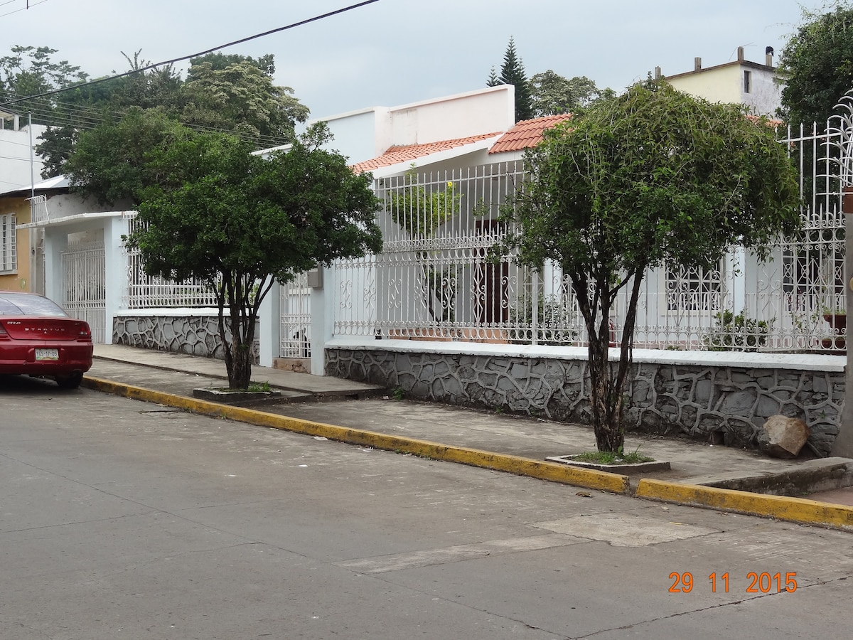 A quiet street scene is depicted, featuring two neatly trimmed trees along a stone wall. A white residence with a red-tiled roof is visible behind a white metal fence, providing a sense of openness and security. The pavement is clean and well-maintained.