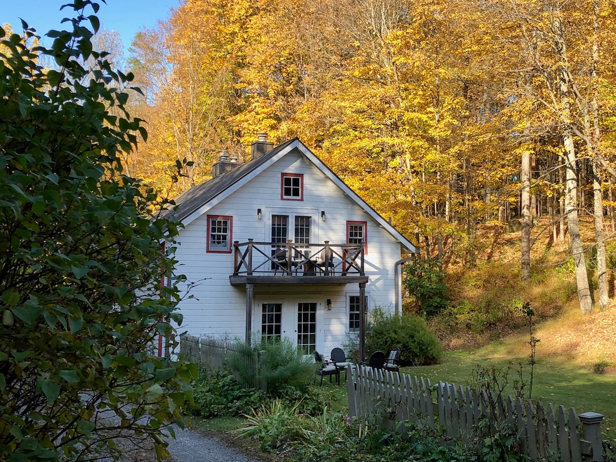 The exterior of a charming two-story house is surrounded by trees adorned with vibrant autumn leaves. A small balcony is visible on the second floor, with red-trimmed windows providing a warm contrast to the white walls. Greenery and a simple fence frame the landscaped yard.