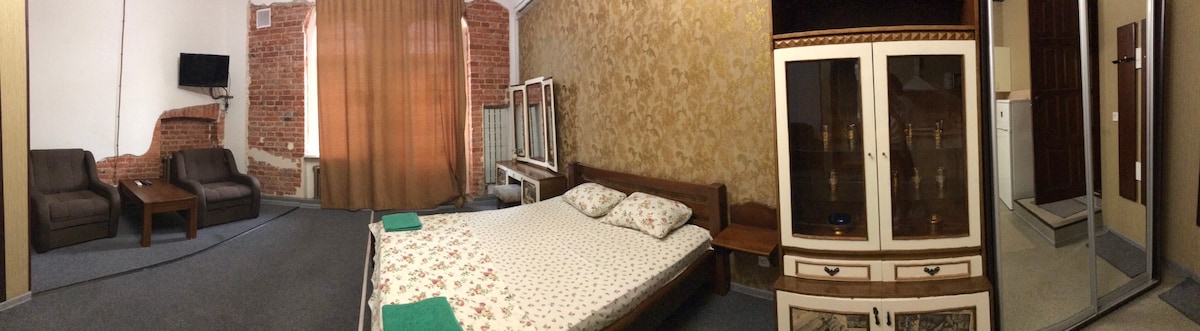 A spacious room is depicted with a large bed featuring a floral-patterned duvet and two pillows. Two brown chairs and a wooden coffee table are positioned near a window draped with light brown curtains. A television is mounted on the wall, and a separate bathroom area is visible.