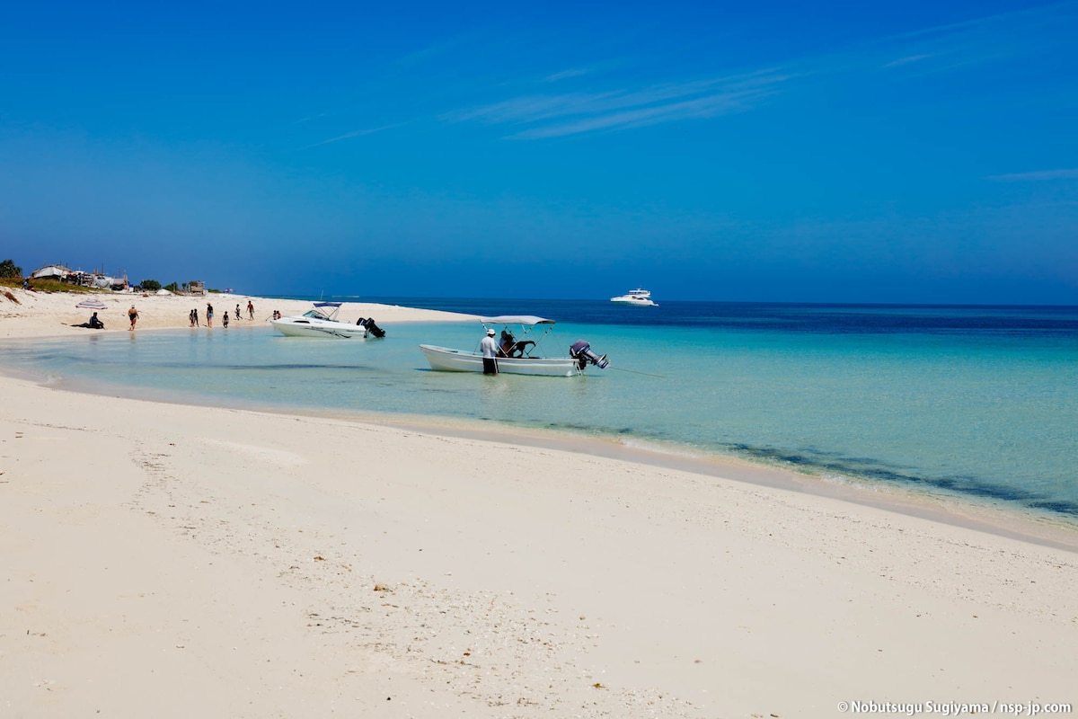 A serene beach scene features soft, white sand curving along a gentle shoreline. A few boats are anchored in clear, turquoise waters. The sky is bright and blue, with distant figures walking along the beach, enjoying the tranquil setting.