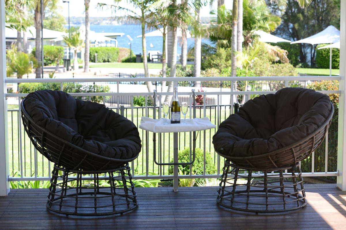 Two oversized, round chairs are positioned on a balcony, overlooking a serene lake. A small table between the chairs is set with two glasses and a bottle. Lush greenery and poolside cabanas are visible in the background.