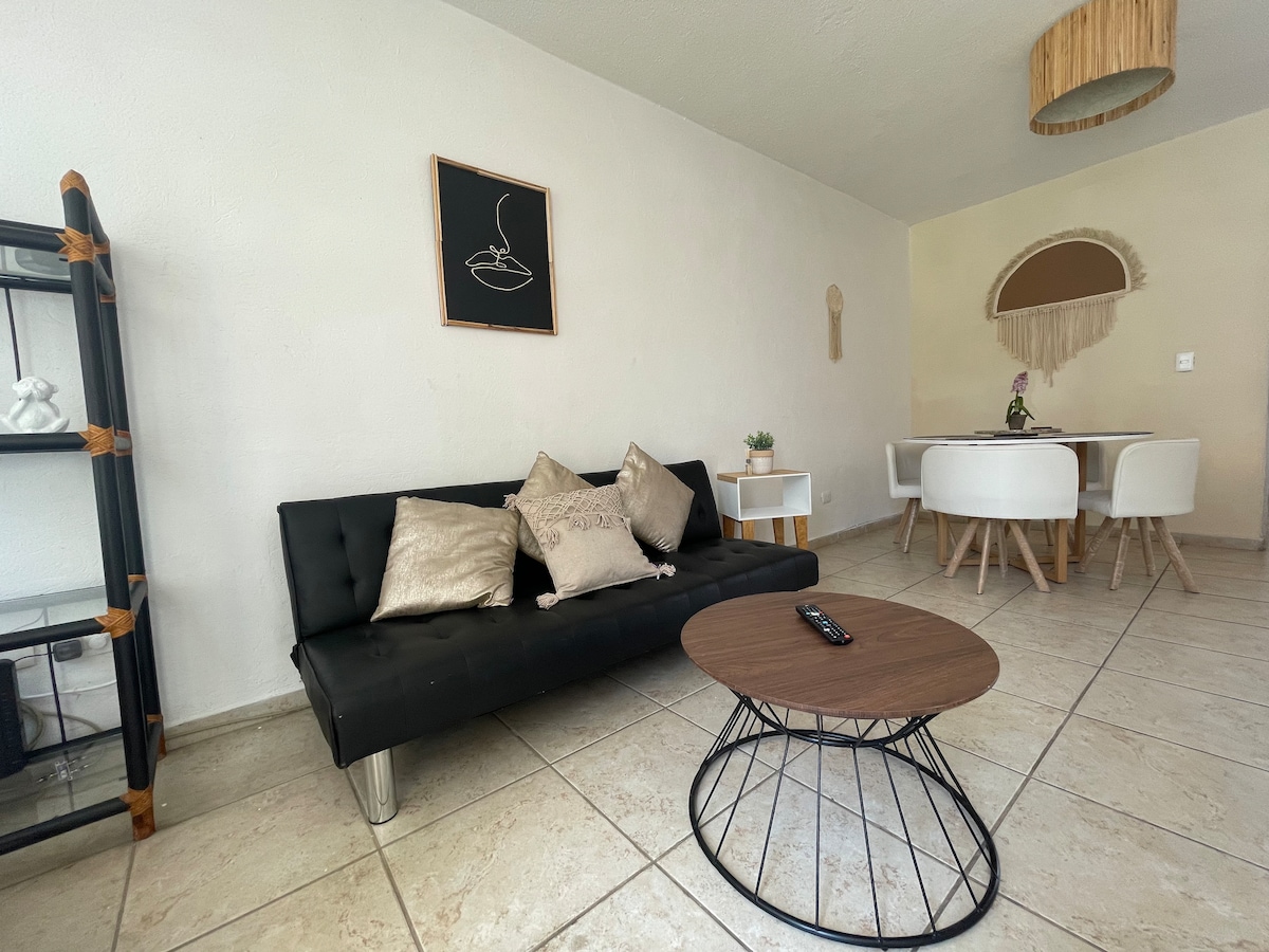 A comfortable living area features a sleek black sofa adorned with decorative pillows. A round wooden coffee table sits in front of the sofa. In the background, a dining space is highlighted, showcasing white chairs around a minimalist table.