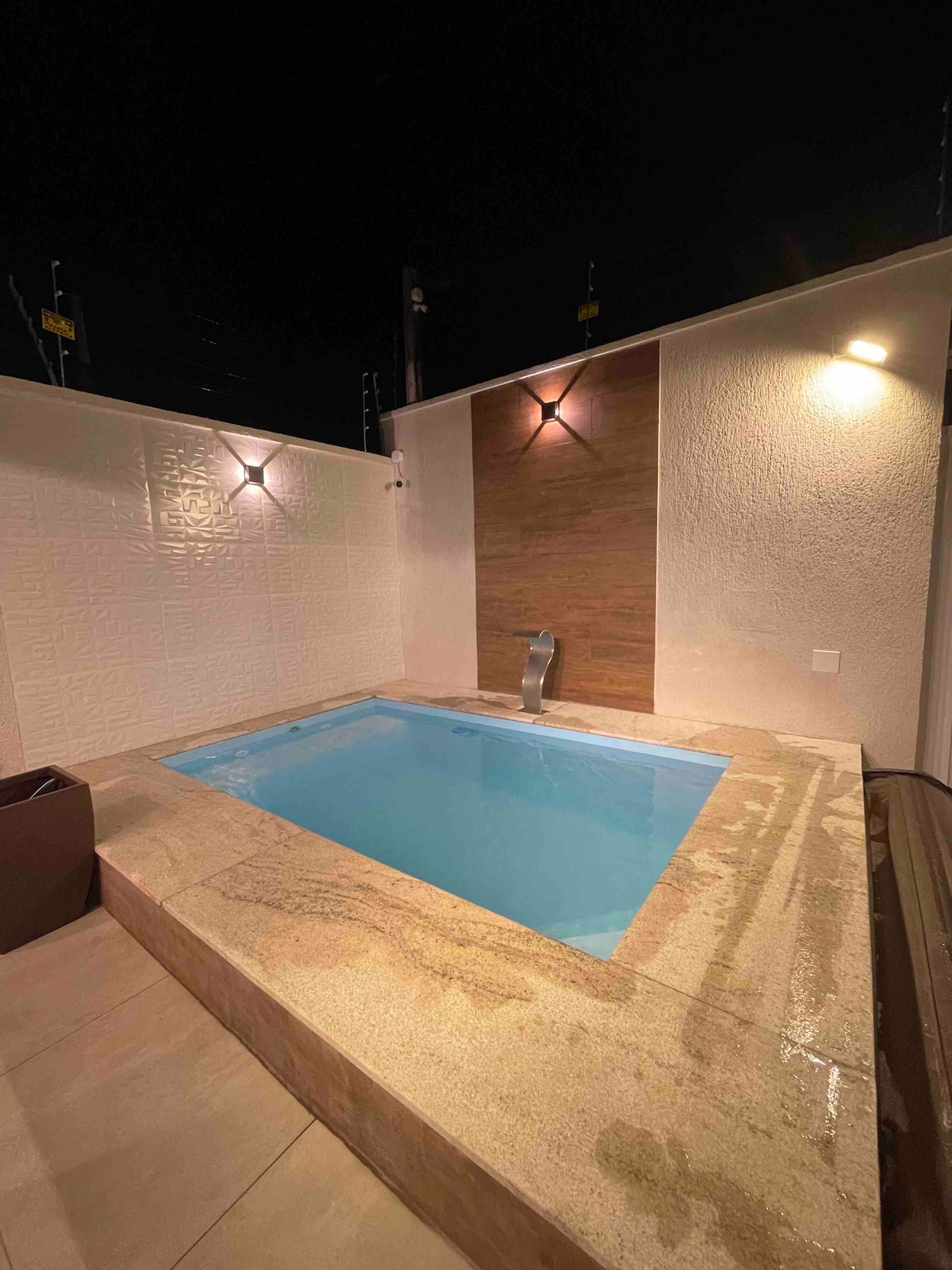 A compact, serene spa area features a small plunge pool with clear blue water. Soft lighting illuminates the surrounding walls, highlighting the textured surface. A water feature is positioned on one side, adding a calming element to the space.