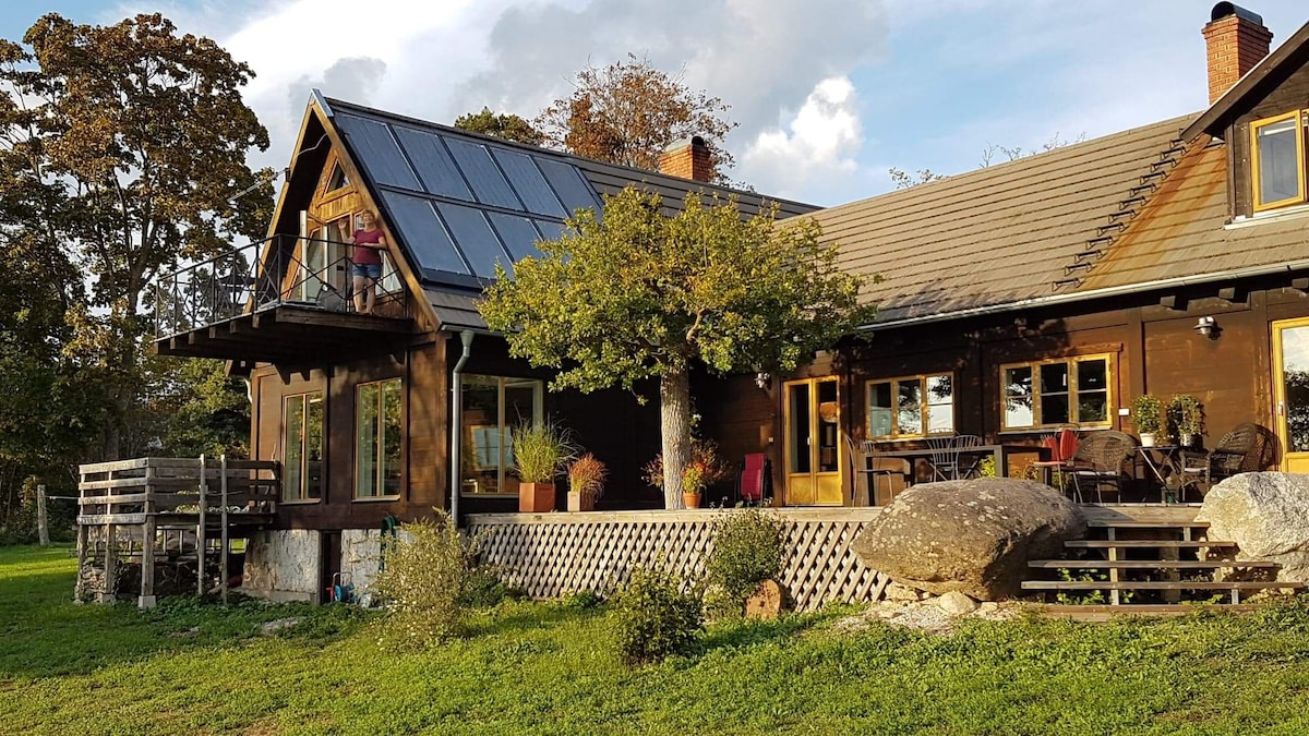 A rustic house is depicted with a large roof featuring solar panels. The structure includes a spacious porch adorned with seating for relaxation. Surrounding greenery and well-placed rocks enhance the natural setting, while trees provide dappled sunlight across the yard.