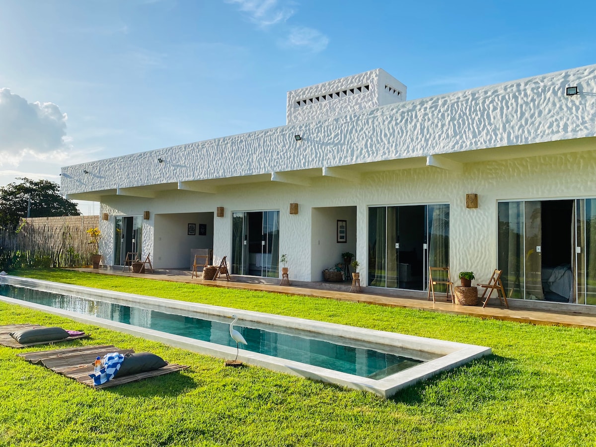 A modern structure features a white exterior and expansive glass doors that open toward a manicured lawn. A long pool runs parallel to the building, surrounded by cozy seating areas, and pots of greenery. The sky is bright blue with hints of soft clouds.