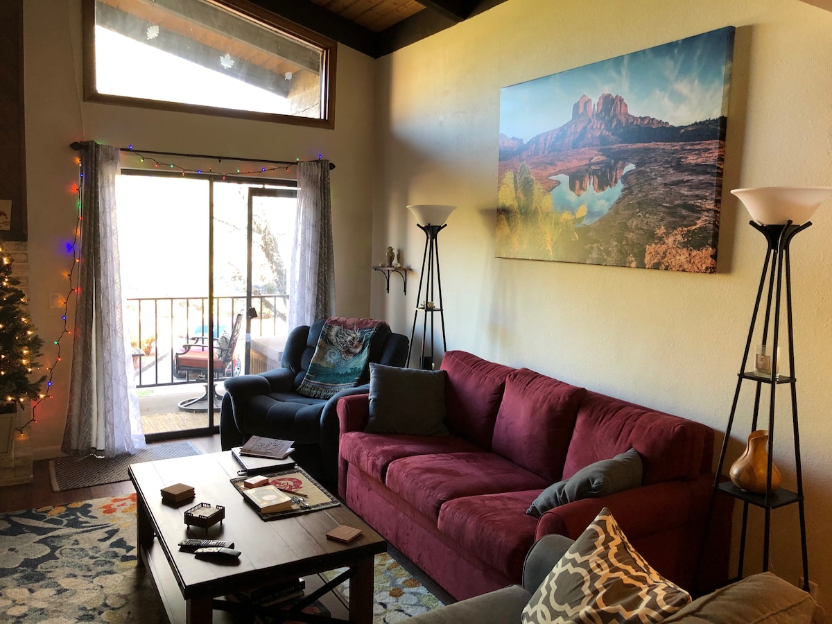 A spacious living area is presented with a deep red sofa, a wooden coffee table, and standing lamps. A large window offers natural light and views of the outdoors. A striking landscape photograph adorns the wall, contributing to a relaxing environment.