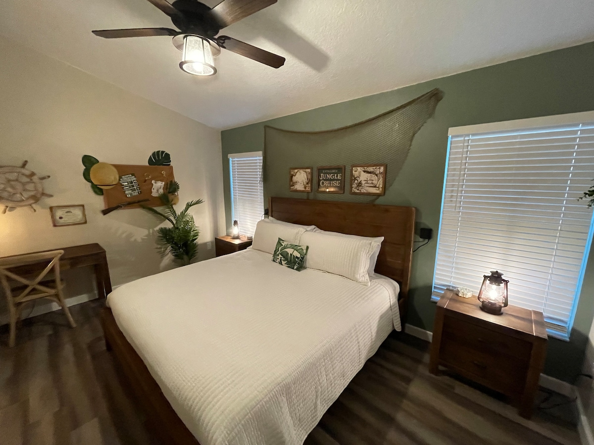 A well-designed bedroom features a spacious bed with white linens and decorative cushions. Natural light enters through two windows, complemented by a small desk and a fern. Lanterns provide soft lighting, while subtle nautical decor accents the green-painted wall.