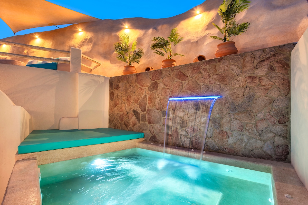The private jacuzzi features a gentle water fountain against a stone wall, creating a soothing atmosphere. Soft lighting enhances the space, while potted plants add a touch of greenery. A comfortable area for relaxation is available beside the jacuzzi for enjoyment.