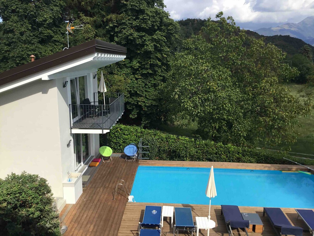 A modern villa is set against a backdrop of lush greenery, with a serene swimming pool surrounded by sun loungers. A balcony extends from the upper level, offering views of the landscape and pool area below. A parasol provides shade along the poolside.