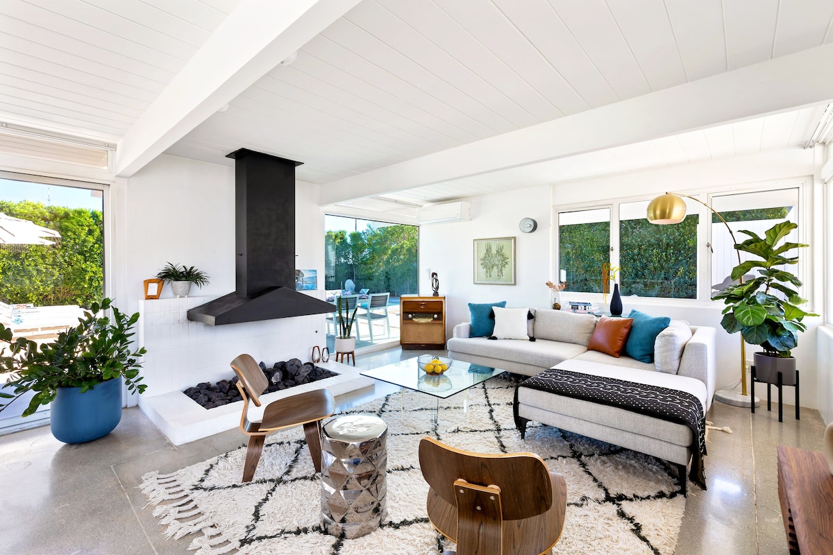 The spacious living area is complemented by large windows that offer views of the outdoor space. A mid-century modern sectional sofa is accented with decorative pillows, while a unique fireplace serves as a focal point. Indoor plants add a touch of greenery to the warm ambiance.