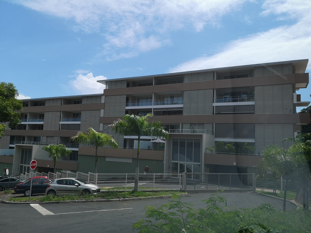 The modern apartment building features multiple floors with large balconies and expansive windows. Tropical plants are positioned near the entrance, which is accessible from a landscaped path. Several parked cars are visible in front of the building, with a clear blue sky overhead.