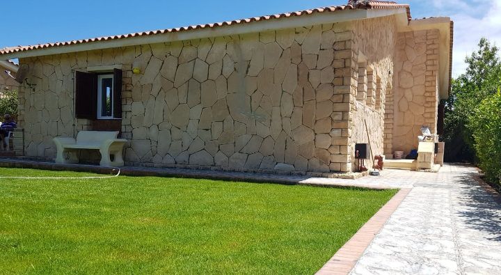 A single-story chalet is showcased with a natural stone facade, complemented by a manicured lawn. A stone pathway leads to a shaded terrace area, and a small garden is visible to the side, bordered by shrubs and greenery.