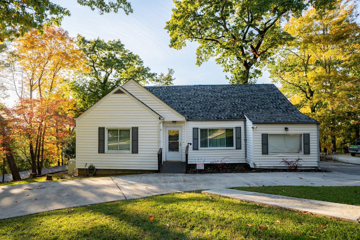 The exterior of the house features a well-maintained white facade with gray shutters, surrounded by vibrant autumn foliage. A spacious driveway leads to the entrance, framed by green grass and a few mature trees. Natural light enhances the welcoming appearance of the home.