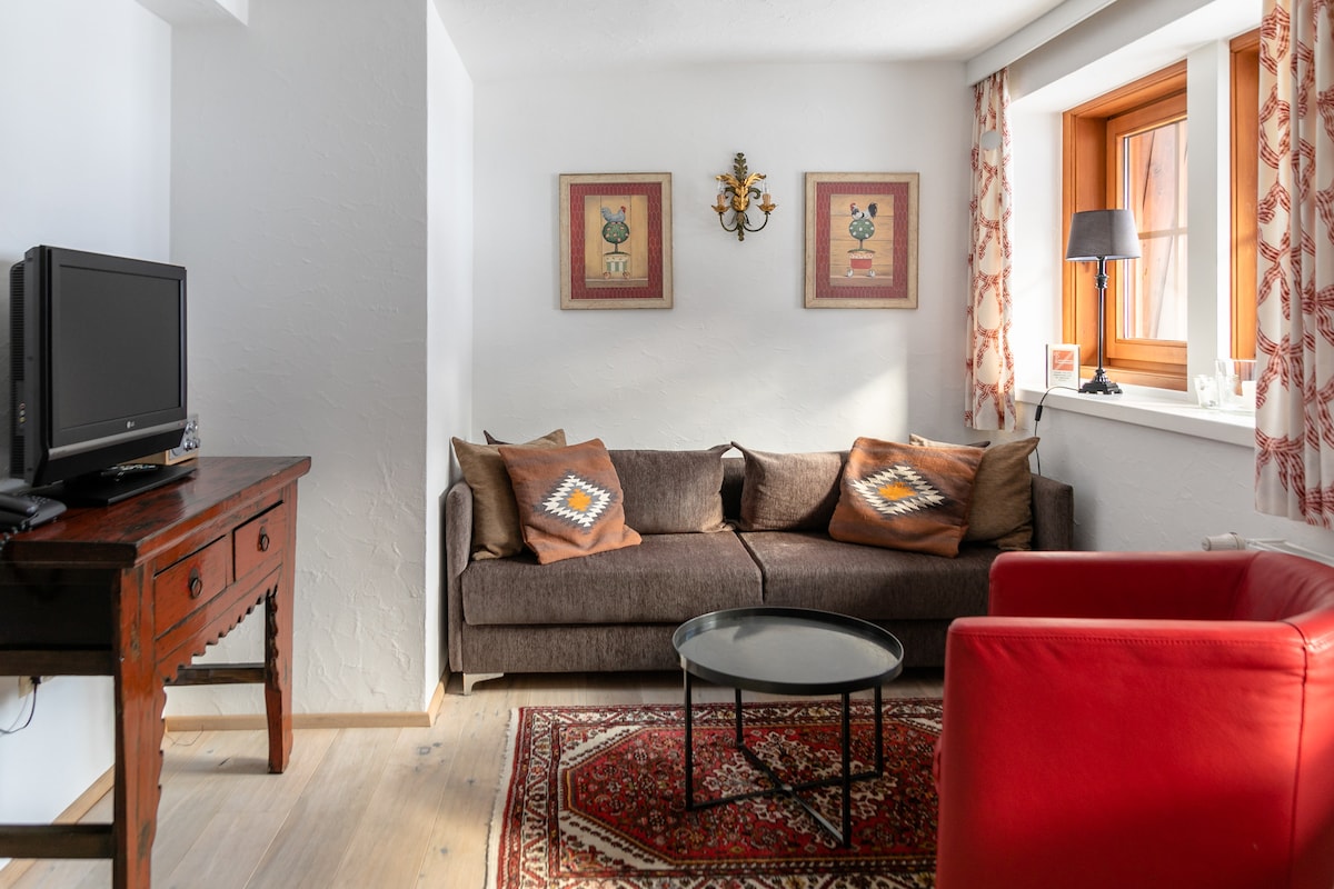 An inviting living area features a brown sofa adorned with decorative cushions opposite a small round table. A rich area rug lays on the light wooden floor, enhancing the space. Two framed artworks and a lamp by the window add visual interest.