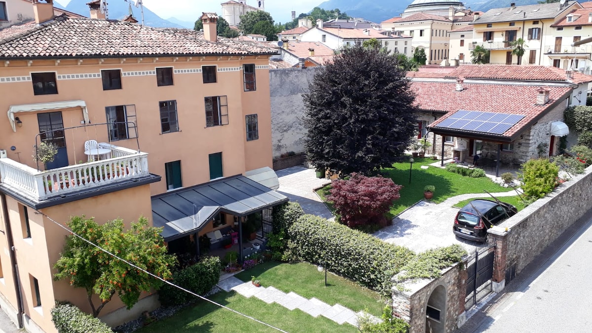 An aerial view showcases the well-maintained outdoor spaces, featuring a green lawn and a well-kept garden with shrubs and a tree. The building's warm-colored exterior is complemented by a shaded porch and solar panels on the adjacent structure, situated within a tranquil area.