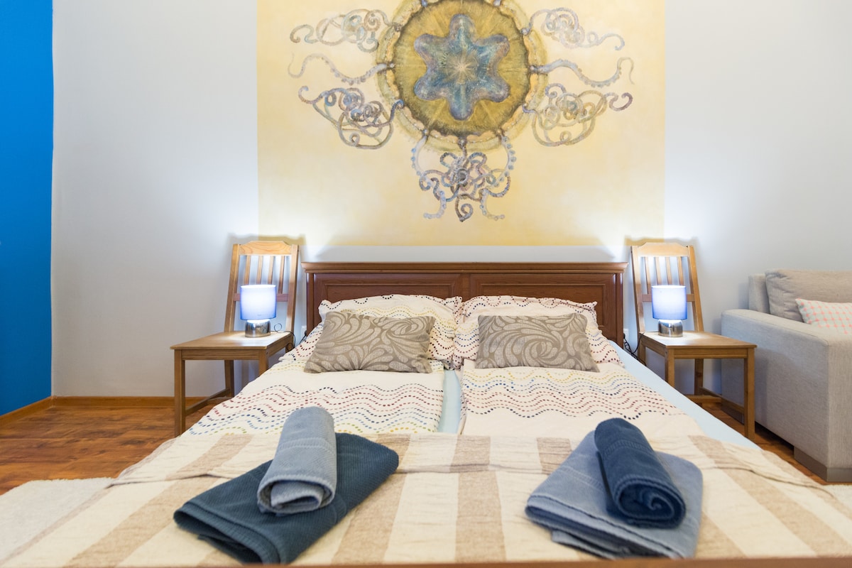 A spacious bedroom is featured, with a large bed adorned with a textured blanket. Night tables with lamps are positioned on either side, while artwork above the headboard adds character. A sofa is visible in the corner, creating a cozy atmosphere.