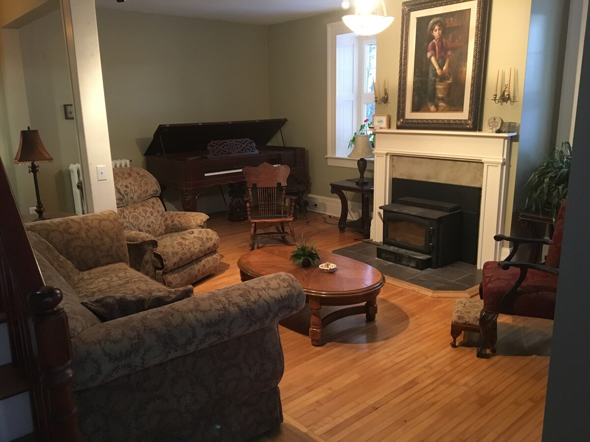 The living room features a warm wooden floor and is furnished with a large patterned couch, a cozy armchair, and a rocking chair. A circular wooden coffee table sits in the center, while a fireplace and a piano are positioned against the wall. Natural light filters in through the window.