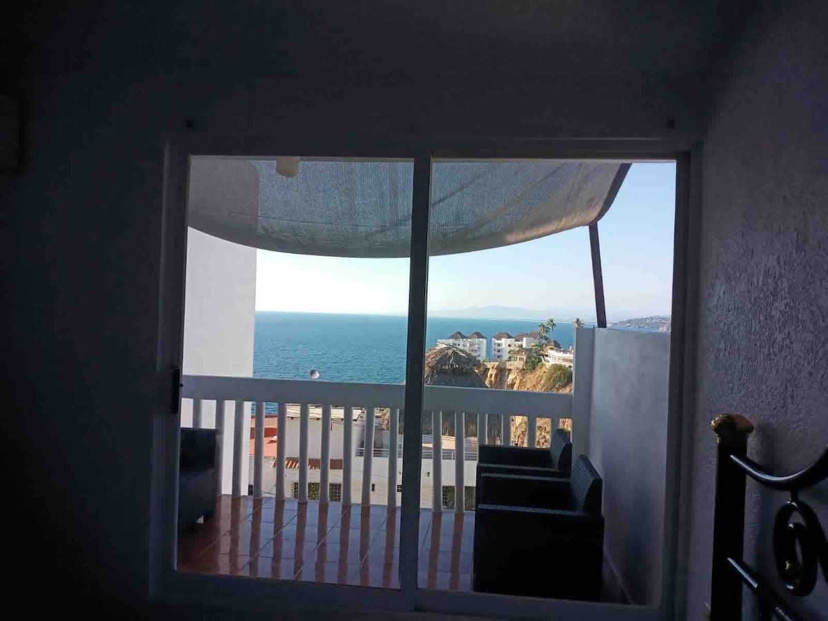 A view is captured through large glass sliding doors, revealing a serene ocean landscape. A shaded balcony is visible, with two dark chairs placed on a tiled floor. The distant shoreline and mountains frame the tranquil waters, enhancing the sense of open space.