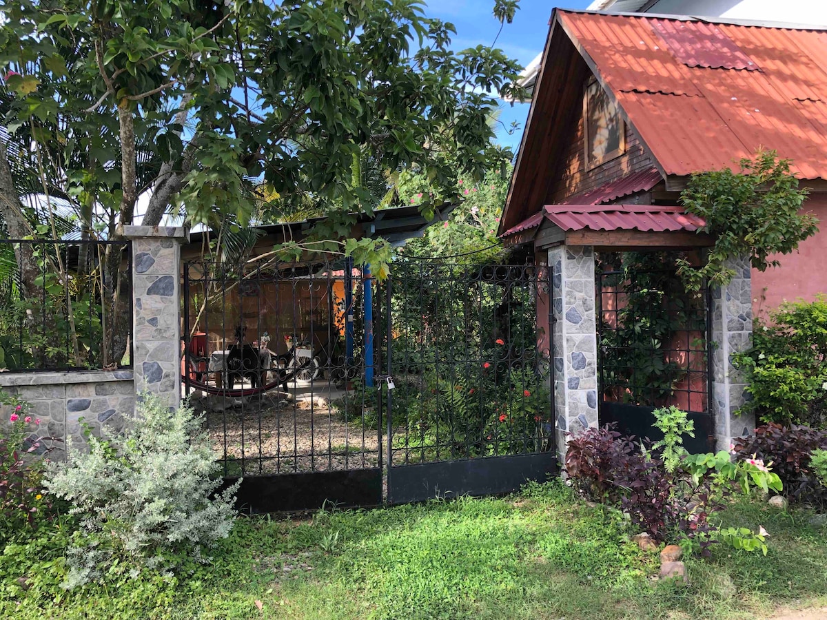An inviting entrance is framed by stone pillars and a wrought iron gate. Lush plants and flowers surround the entryway, leading to a partially visible outdoor space with seating. A rustic roof adds character to the nearby structures, set in a serene green landscape.