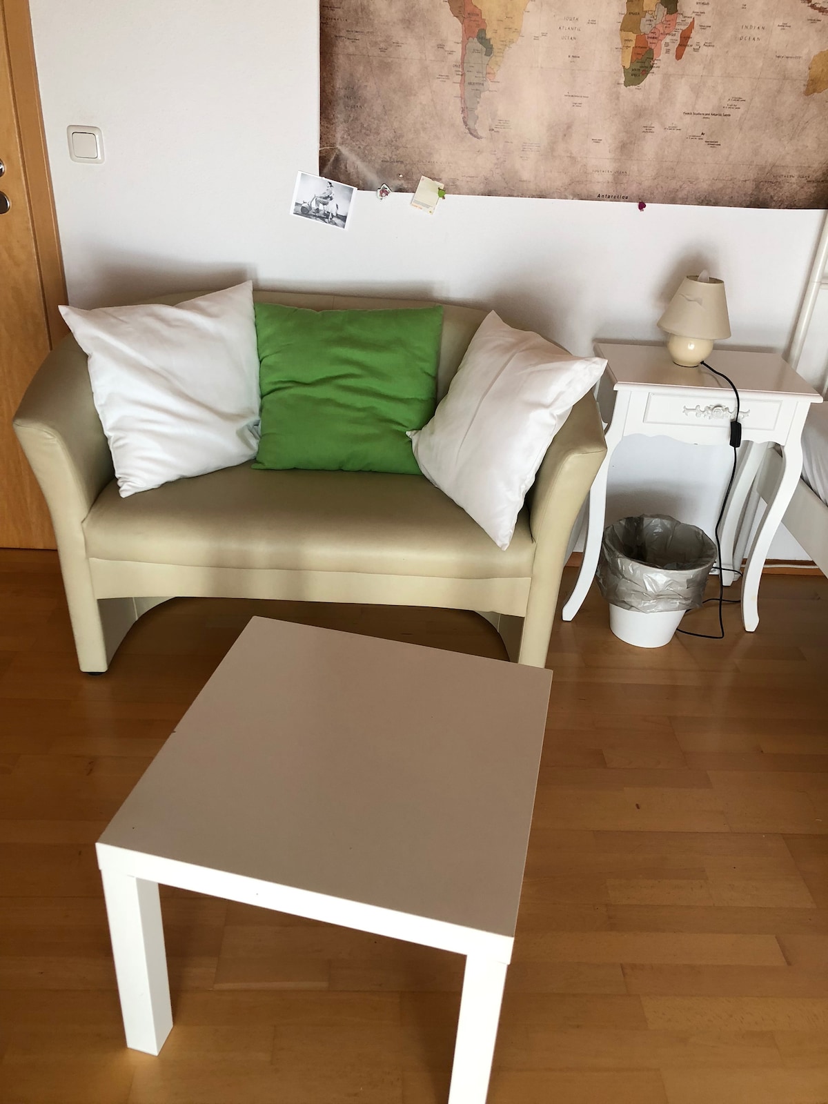 A cozy seating area includes a light-colored armchair adorned with white and green pillows. A small white table is positioned in front of the chair, while a bedside lamp and a trash bin are placed beside the chair. A world map decorates the wall, adding a global touch.