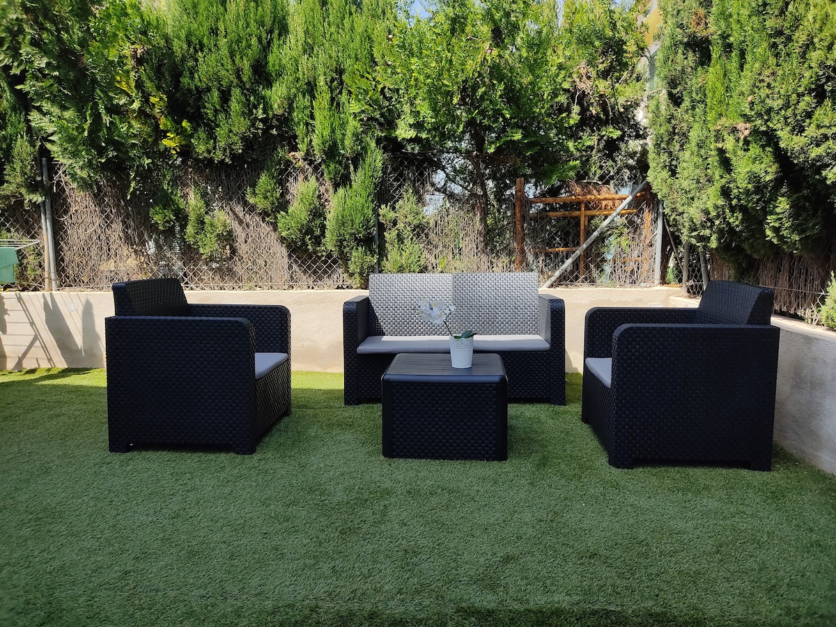 An outdoor relaxation area is furnished with a set of four modern wicker chairs and a matching coffee table. The green artificial grass is surrounded by tall trees, providing a tranquil setting for guests to unwind.