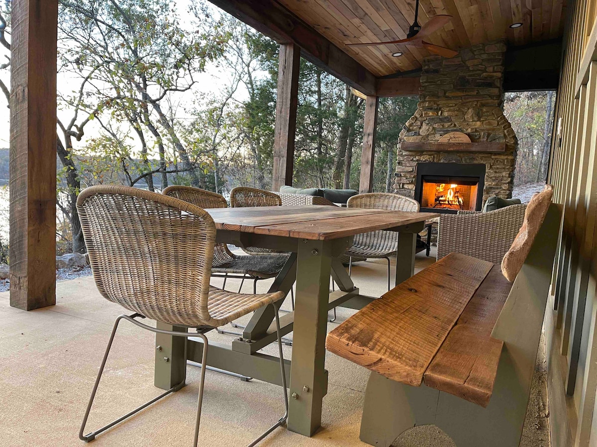 An outdoor dining area is showcased under a wooden overhang, featuring a large wooden table surrounded by woven chairs. A stone fireplace is positioned in the corner, providing warmth. Natural light filters through the trees, highlighting the serene lakeside setting.