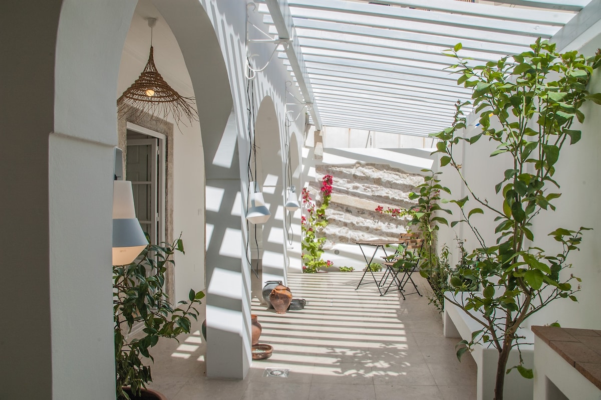 A central private patio is revealed, featuring white arches and a slatted roof allowing dappled sunlight to filter through. Potted plants and a small table with chairs are placed among the greenery, creating a serene outdoor space suitable for relaxation.