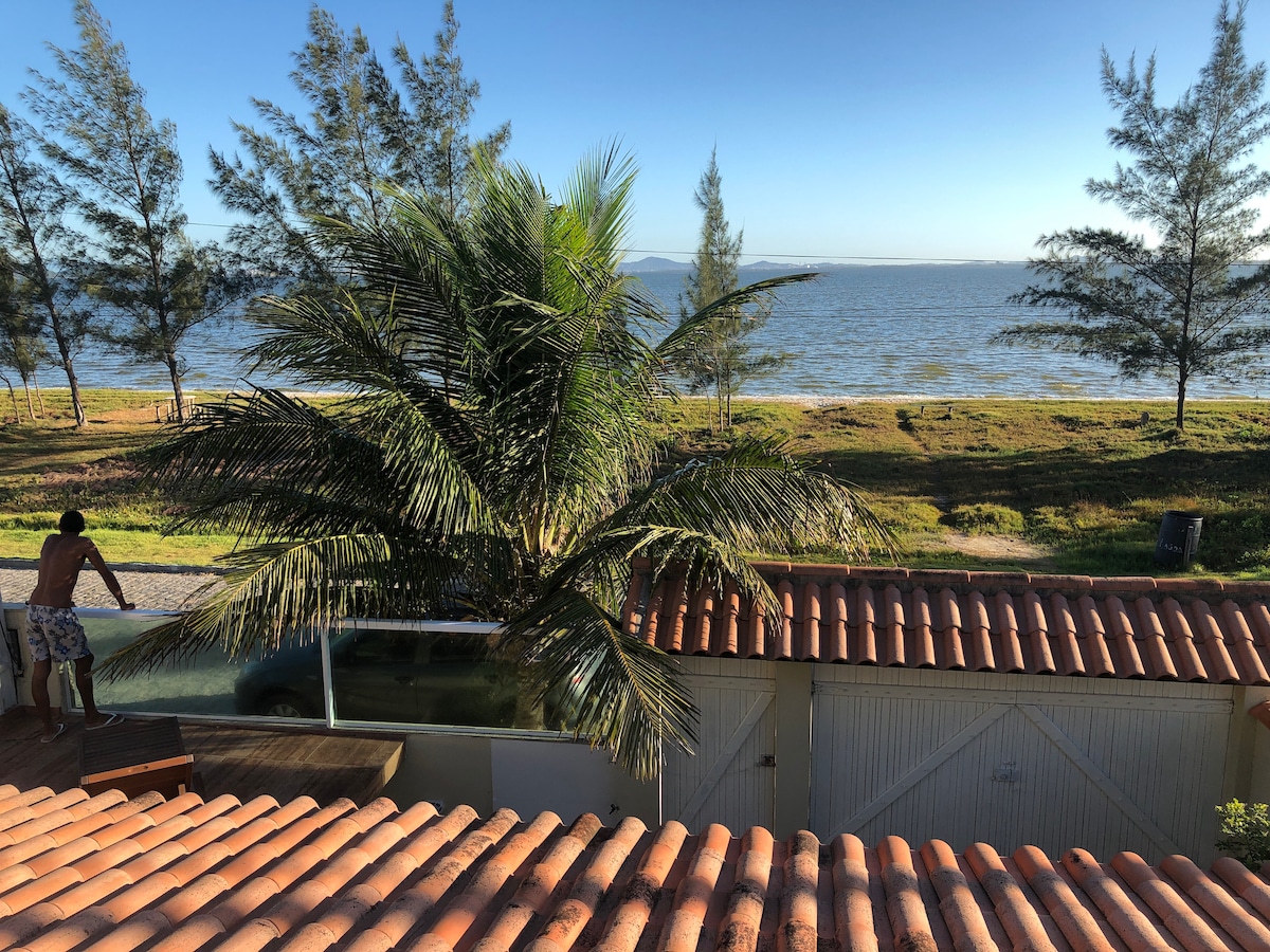 A view of the tranquil beach can be seen through a lush palm tree. The shoreline is adorned with gentle waves under a clear blue sky, while terracotta rooftops frame the picturesque scene from above.