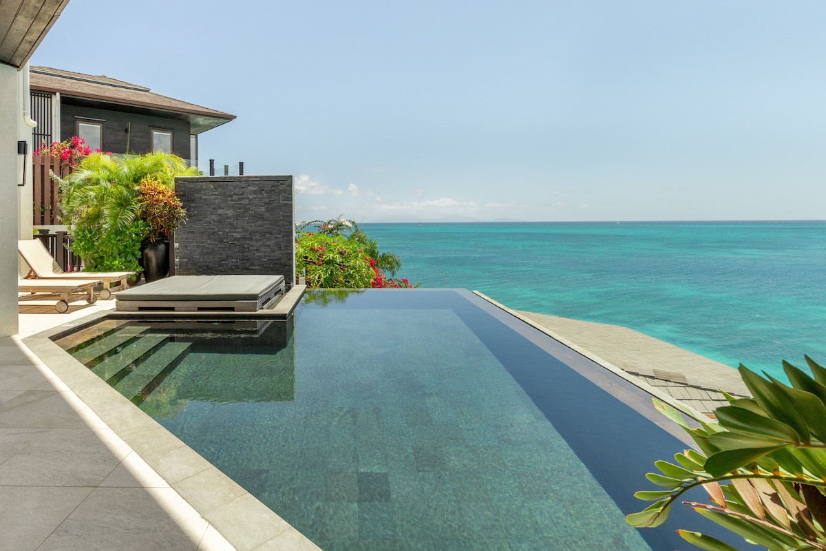 An infinity pool extends towards the ocean, surrounded by lush greenery. The pool's edge creates a seamless transition to the water, reflecting the clear blue sky. Sun loungers are positioned nearby, providing a relaxing space to enjoy ocean views.