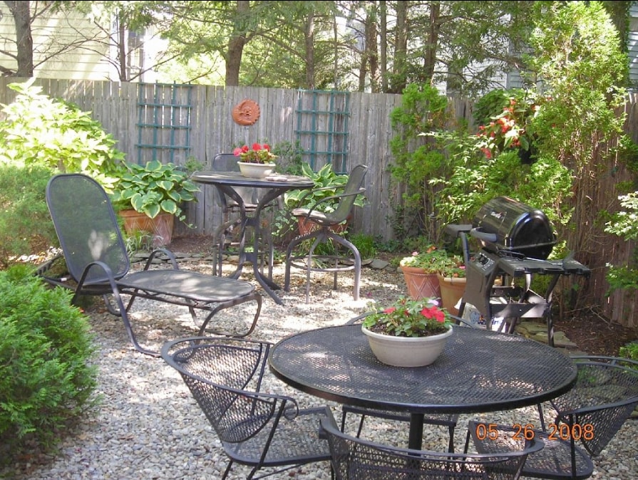A landscaped private yard is surrounded by greenery, featuring various seating options including tables and chairs. Potted flowers add color, while a gas grill is positioned for outdoor cooking. The space is designed for relaxation and gatherings among friends.