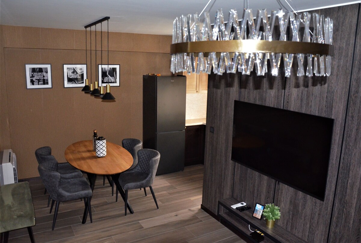 A contemporary dining area features a round wooden table surrounded by six upholstered chairs. A stylish light fixture hangs overhead. A flat-screen TV is mounted on the wall beside decorative photos, and a sleek kitchenette is visible in the background, enhancing the modern design.