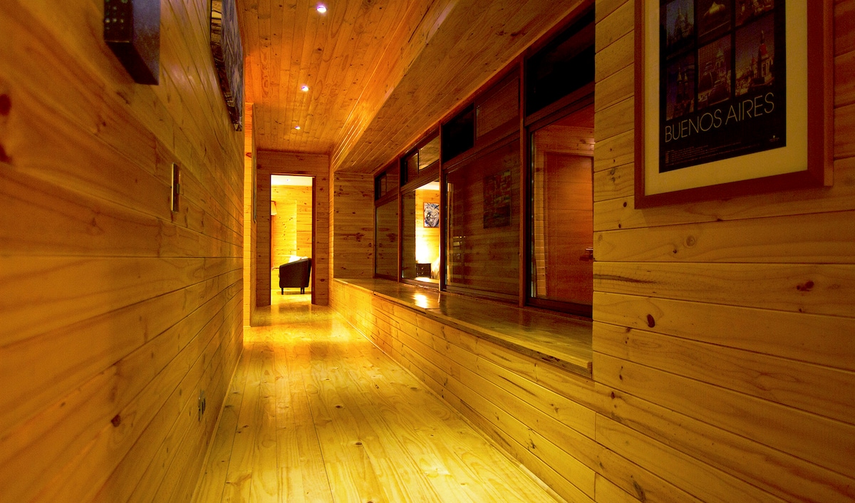 A long hallway features wooden walls and flooring, creating a warm and natural ambiance. Windows line one side, allowing for natural light to illuminate the space. Artwork adorns the walls, contributing to the modern design of the home.