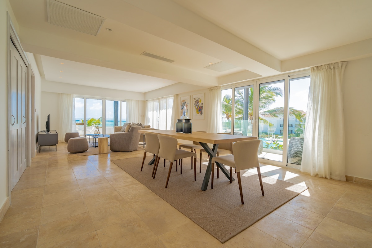 A spacious living and dining area features large glass doors that allow natural light to flood in. A neutral color palette is complemented by modern furnishings, including a dining table with several chairs, a comfortable seating area, and artwork adorning the walls, with views of the exterior tropical landscape.