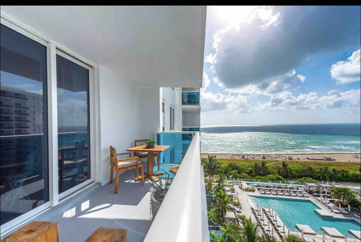 A private balcony features a small wooden table and chair set, overlooking the ocean and pools below. Natural light illuminates the space, while expansive glass doors provide unobstructed views of the Atlantic and the vibrant scene of South Beach.
