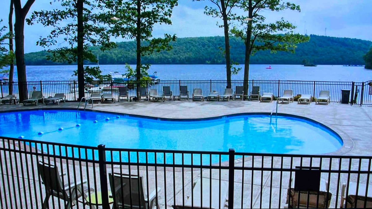 A rectangular pool is framed by a black fence, with lounge chairs arranged along its perimeter. Lush trees provide shade, and a serene lake is visible in the background, enhancing the tranquil setting.