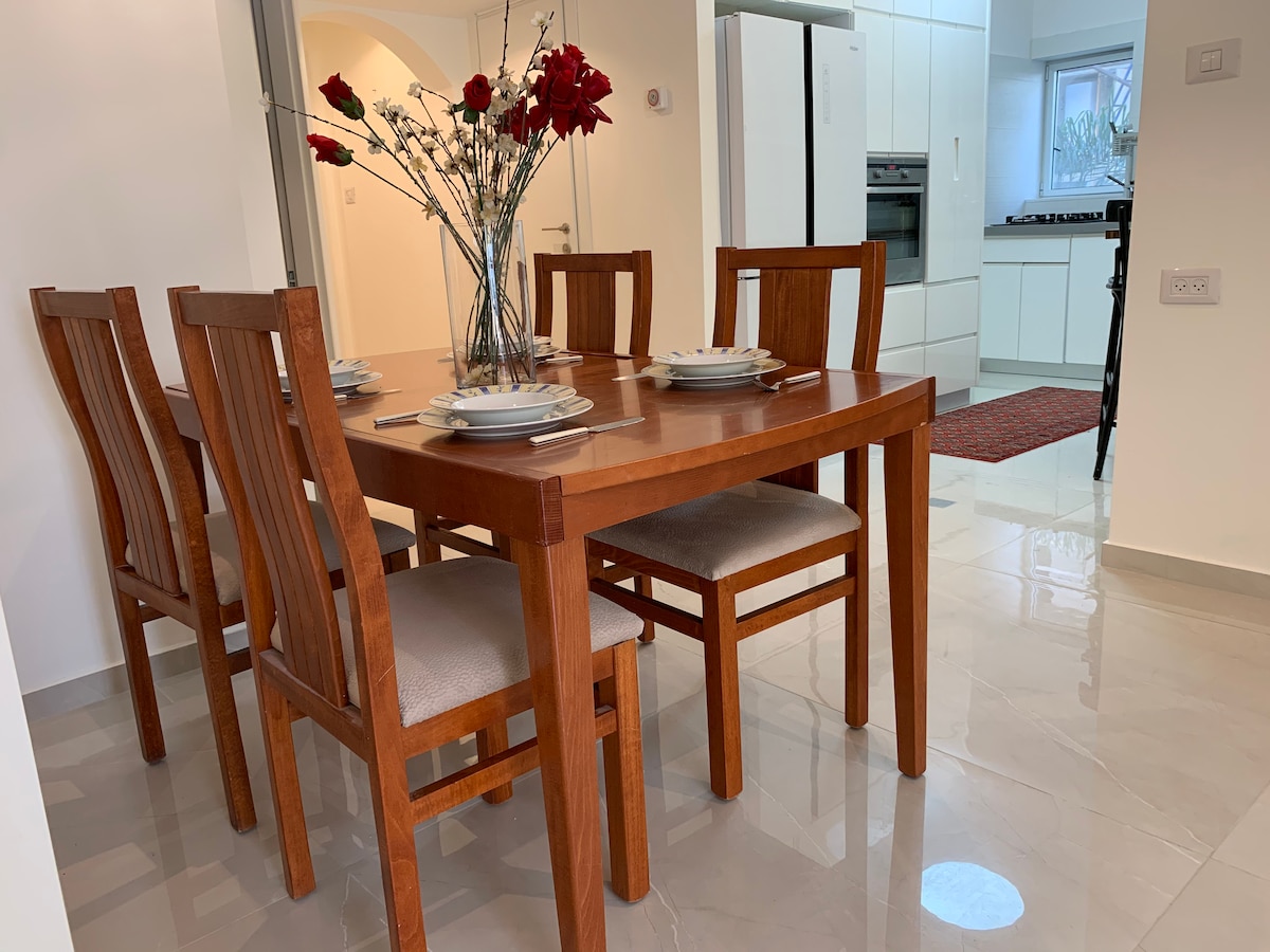 A wooden dining table is set with dishes and cutlery, surrounded by four cushioned chairs. A vase filled with red flowers adds a touch of color. The space features a glossy tiled floor, leading towards a modern kitchen visible in the background.