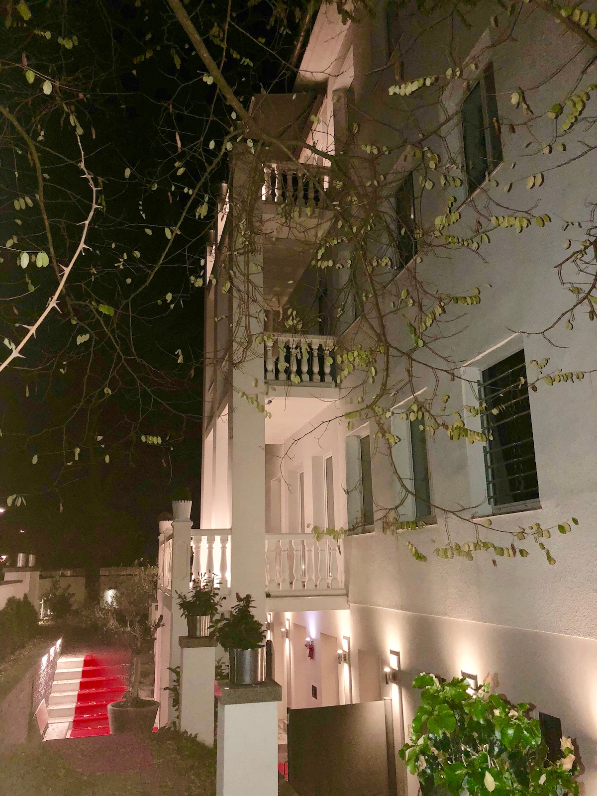 The exterior of a three-story building is illuminated under the night sky, highlighting a stylish façade. Balconies with decorative railings are visible on the upper floors. Soft lighting along the pathway guides towards the entrance, surrounded by greenery.