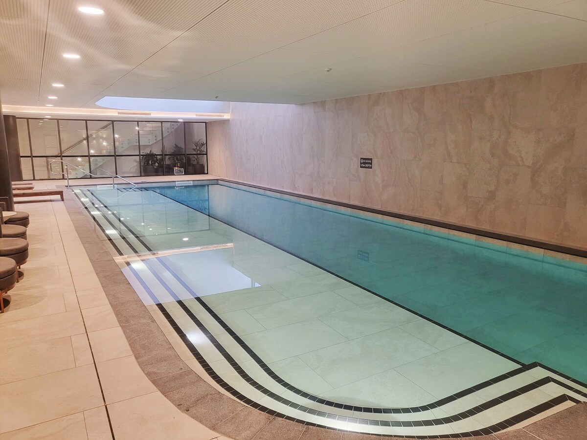 An indoor swimming pool is featured with a smooth tiled surface, illuminated by soft lighting. The pool area is flanked by comfortable lounge seating. Floor-to-ceiling windows allow natural light to fill the space, enhancing the serene ambiance.