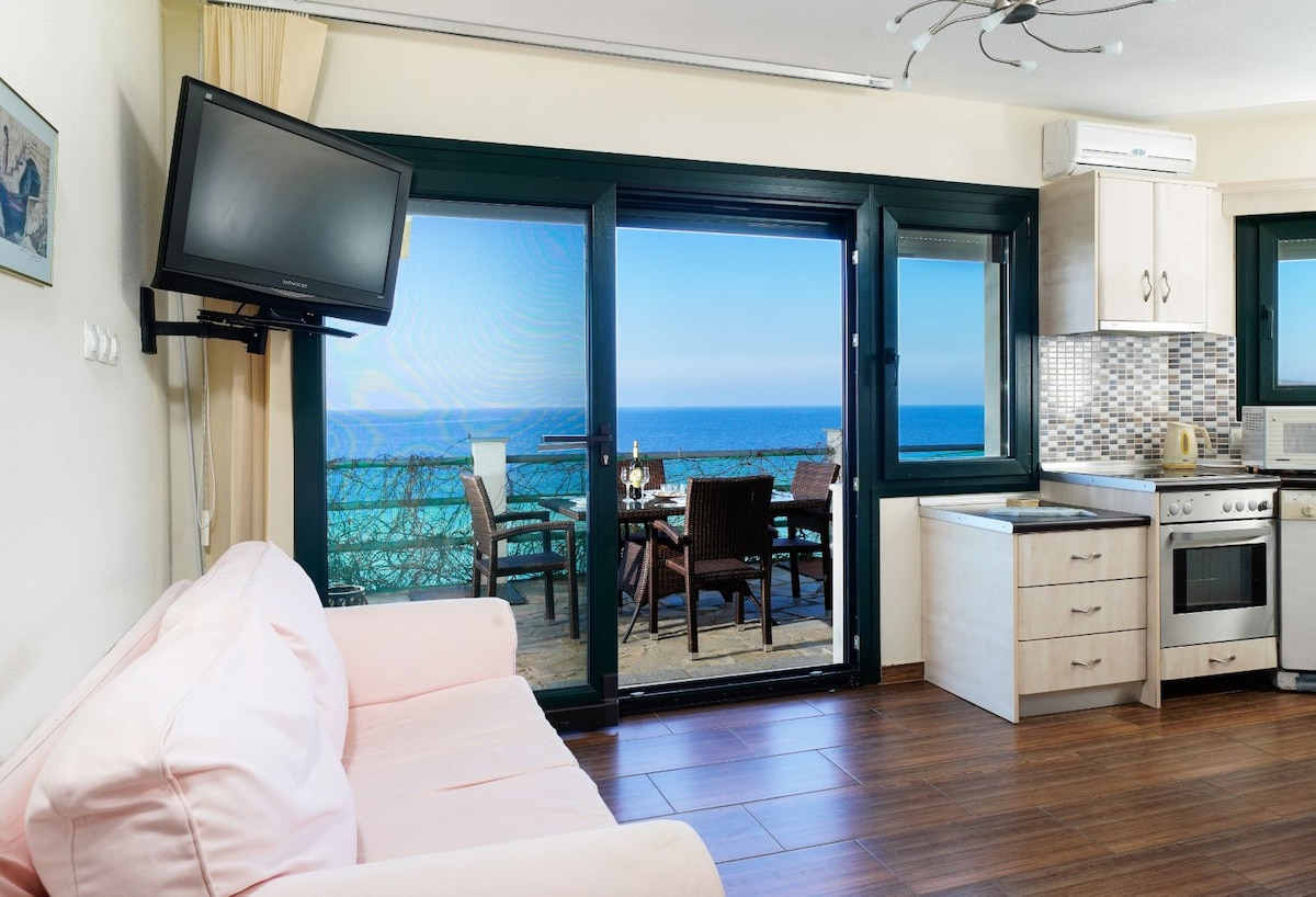 A living space features a soft pink sofa facing large glass doors, revealing a stunning sea view. The open kitchen, equipped with modern appliances, is situated beside the doors. A dining area on the terrace showcases an outdoor table and chairs with glimpses of the ocean.