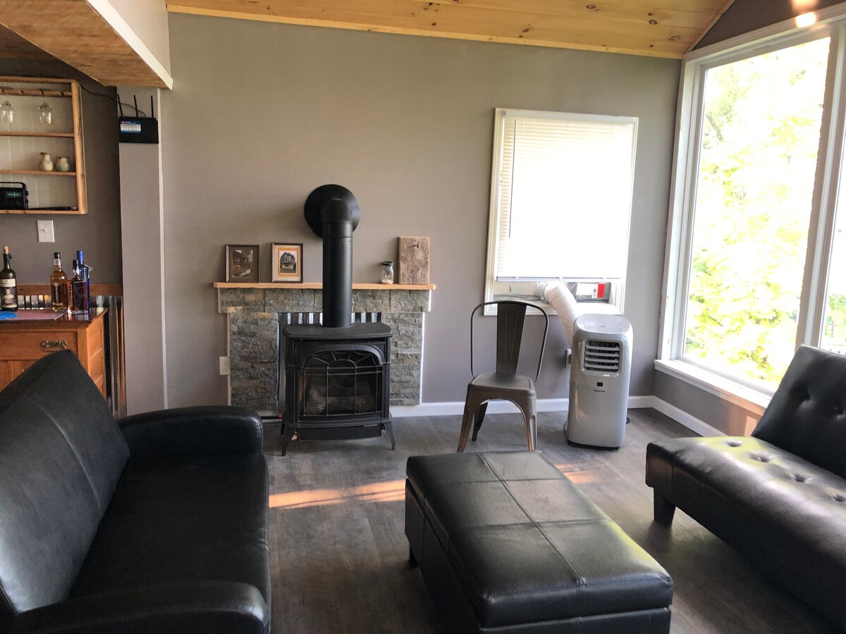 A cozy living area features a custom stone gas stove surrounded by a black couch and ottoman. Large windows provide views of nature, while a stylish bar area is set against the wall, displaying glassware. An air conditioning unit is present for comfort.