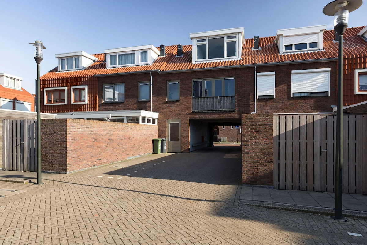 The image showcases the exterior of a modern brick townhome with red tiled roofing. A shared driveway is visible, flanked by wooden fences. The structure features several windows, allowing ample natural light. A covered balcony is positioned on the upper level, adding to the architectural interest.