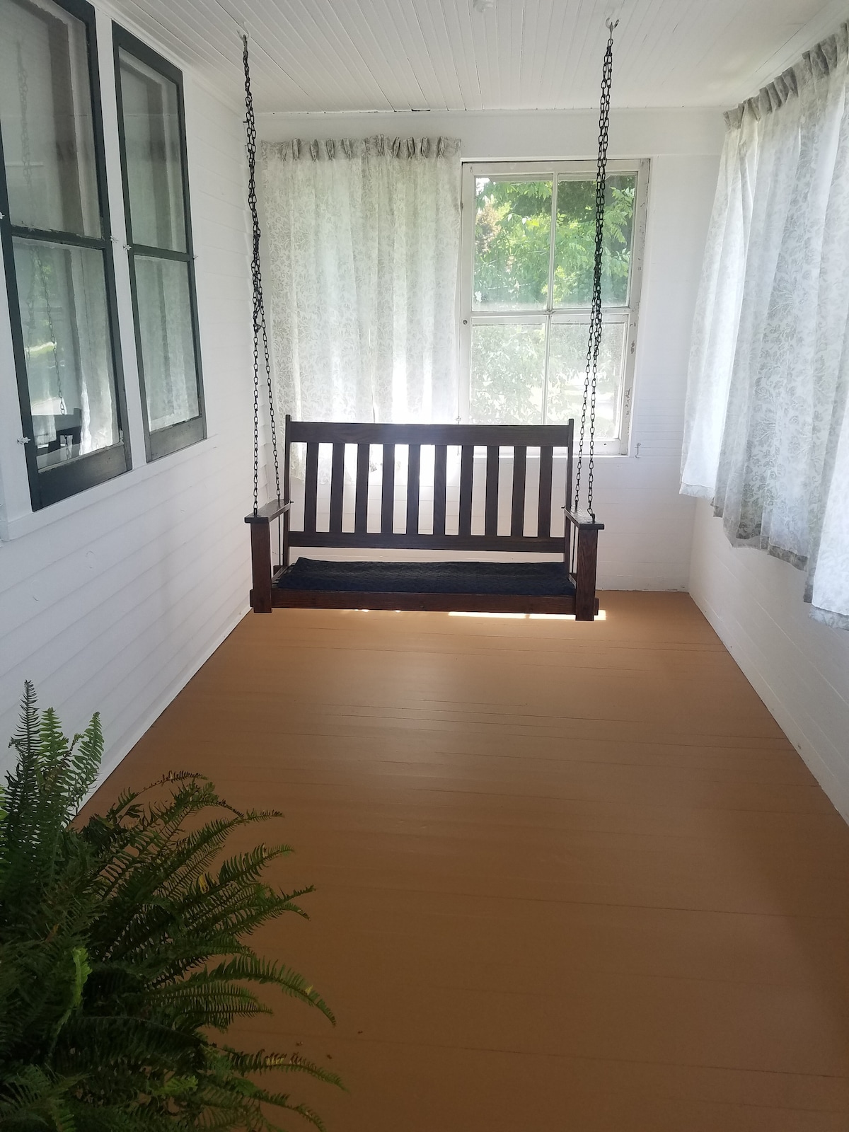 An enclosed three-season porch is shown, featuring a wooden swing bench suspended from the ceiling. Natural light filters through large windows adorned with sheer white curtains. A small fern is positioned on the floor, complementing the warm-toned flooring.