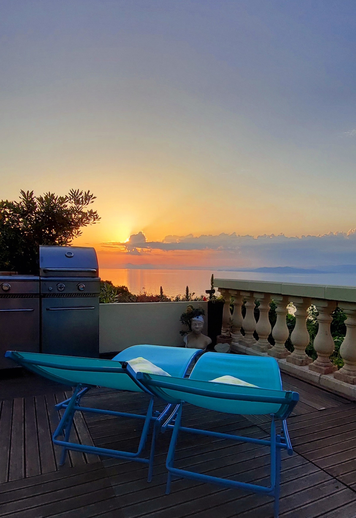 Two turquoise lounge chairs are positioned on a wooden deck, facing a vibrant sunset over the sea. A grill is visible to the left, framed by lush greenery and elegant balustrades that overlook the landscape.