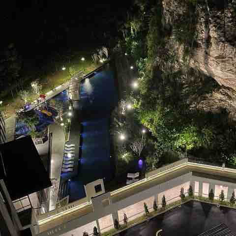 An aerial view captures a serene outdoor pool area illuminated by soft lighting, surrounded by lush greenery and positioned against a natural rock formation. Loungers are placed around the pool, creating a tranquil space for relaxation.