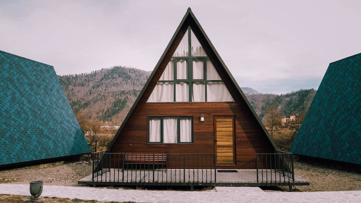 A triangular wooden cottage stands prominently, flanked by two sloped-roof structures. Large windows allow natural light to illuminate the interior. The entrance features a simple railing and a wooden bench, while the surrounding landscape displays rolling hills in the background.