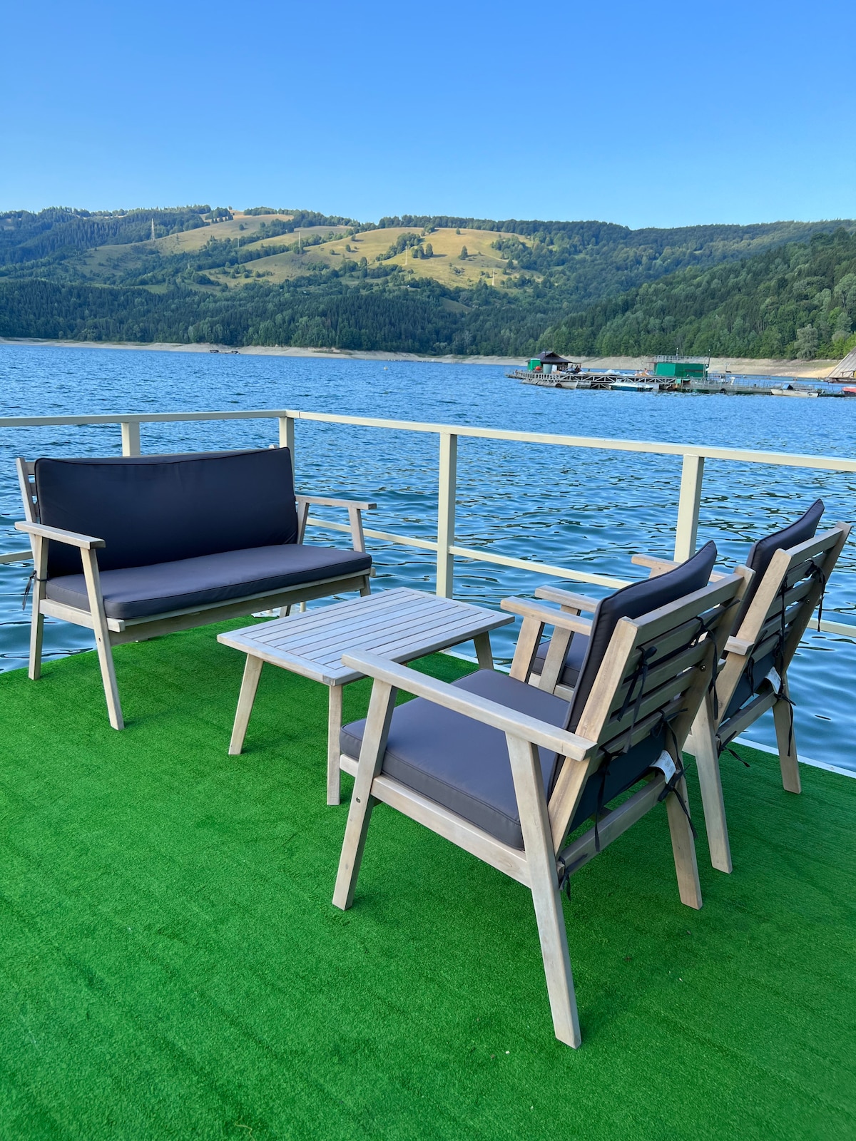 A seating area on the terrace features a gray sofa and two wooden armchairs surrounding a rectangular table. The seat cushions are dark gray, and the floor is covered in green turf. A panoramic view of the lake and mountains is visible in the background.